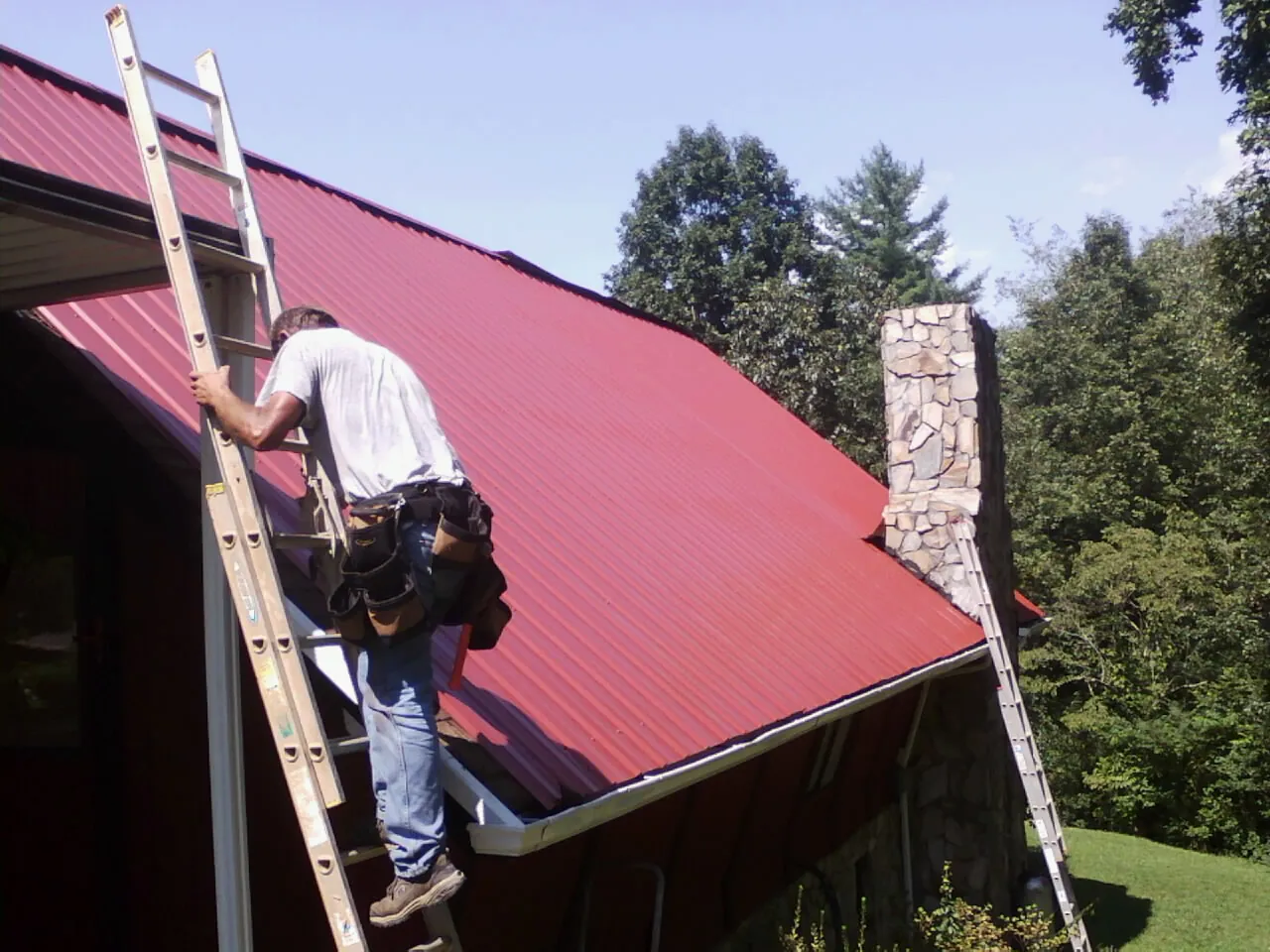Roofer on a ladder beside a red metal roof and stone chimney — Soffit & Fascia Repair by Roofing Brothers in Ellisville